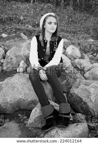 Pretty girl wearing jeans and a varsity jacket sitting on river rocks in black and white