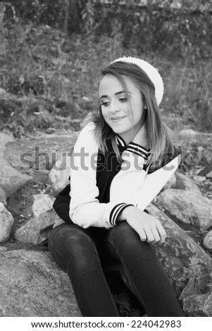 Pretty girl wearing jeans and a varsity jacket sitting on river rocks in black and white