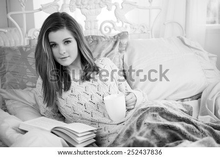 Young woman at home laying in bed reading a book with a cup of coffee in black and white