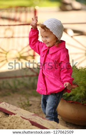 the girl in a pink raincoat stands near a sandbox and waves a hand
