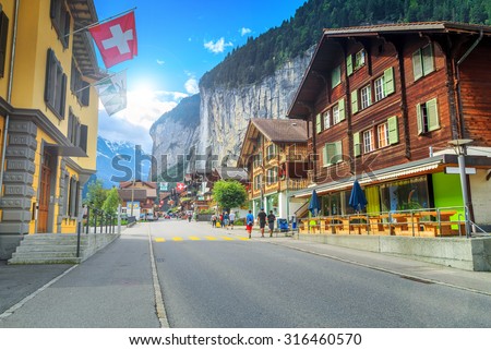 Principal street of Lauterbrunnen with shops, hotels, terraces, swiss flags and stunning Staubbach waterfall in background, Bernese Oberland, Switzerland, Europe