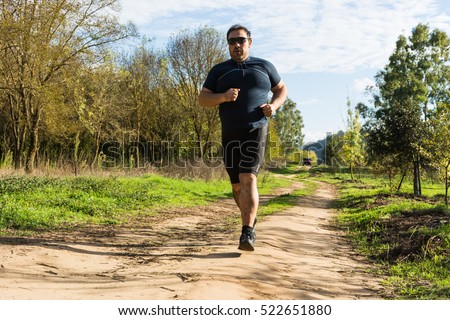 Big belly man jogging , exercising, doing cardio in the park , slightly overweight, loosing weight. On a lawn of green grass between trees without leaves.