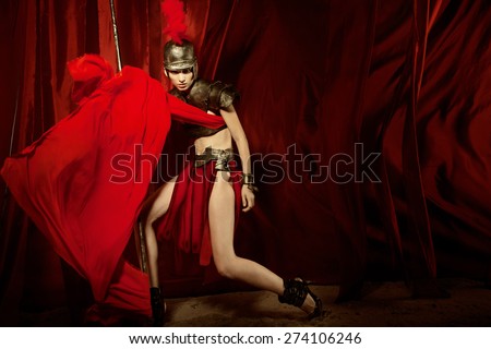 Fashion studio shot of beautiful woman in armour and helmet with feathers holding spear red curtain background