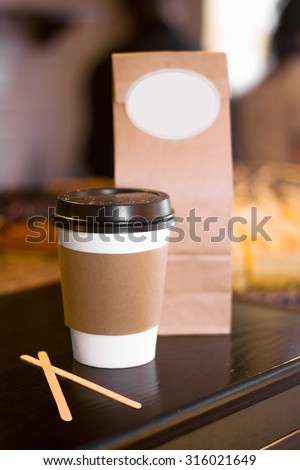 Disposable Paper Cup of Coffee or Tea, covered with black plastic cap and Craft Paper Bag on Wooden Bar or Table with space for logo or design. Classic Coffee Shop Takeaway Modern Concept