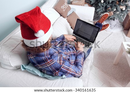Young man with laptop on a sofa near a Christmas tree. Businessman with Santa hat working on the computer in New Years eve.