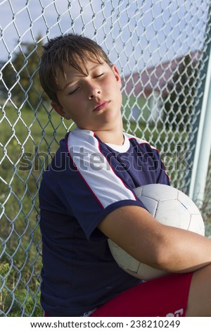 Young football player is tired and resting with the ball at the net.