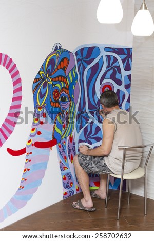 The artist draws a picture sitting on a chair with an elephant on the living room wall.