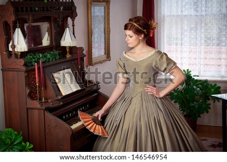 A lovely woman clad in 1860s era historical clothing stands by an old pump organ