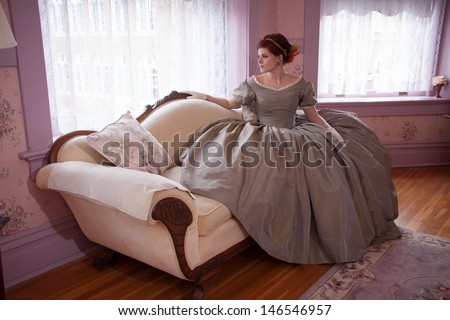 A beautiful woman wearing historical clothing sits on a couch and watches out a window