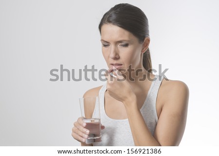 Attractive woman taking medication holding a glass of water in one hand as she slips a tablet or antibiotic into her mouth to treat an illness, or a painkiller or supplement