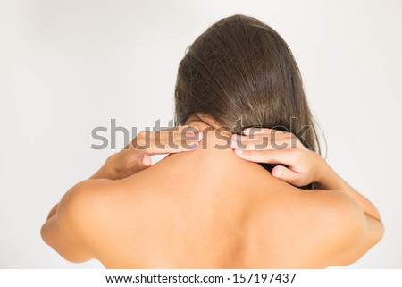 Woman with upper back and neck pain standing naked with her back to the camera and her hand rubbing her shoulder muscles close to the spine