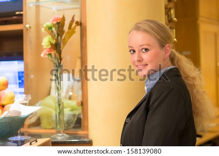 Beautiful stylish blond hotel receptionist standing behind the service desk in a hotel lobby looking at the camera with a friendly smile