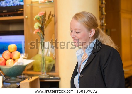 Beautiful stylish blond hotel receptionist standing behind the service desk in a hotel lobby looking at the camera with a friendly smile