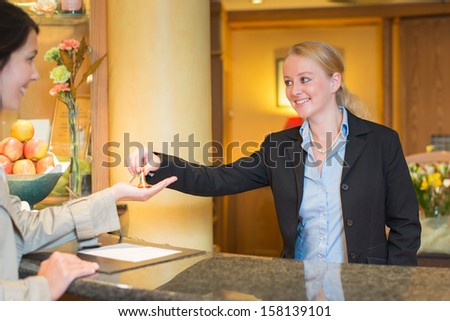 Smiling friendly hotel receptionist standing behind the service desk in a hotel lobby booking in a female client handing her the room keys for her stay during her vacation