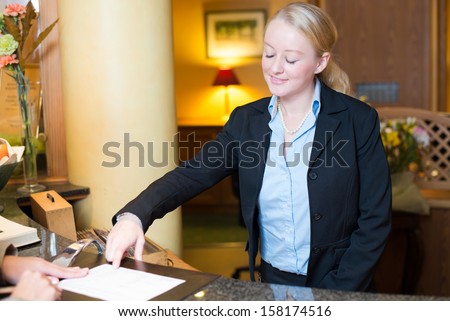 Smiling attractive young receptionist helping a hotel guest check in pointing to information on the form that needs to be completed as they stand at the service desk in the lobby