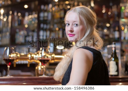 Beautiful young blond woman seated at a bar counter in an upmarket in a hotel, club or restaurant looking back over her shoulder at the camera