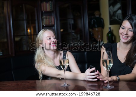 Two beautiful young women enjoying a glass of wine on a night out using their mobile phones laughing at the conversation and text message