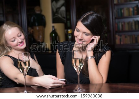 Two beautiful young women enjoying a glass of champagne on a night out using their mobile phones laughing at the conversation and text message