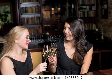 Two beautiful elegant women friends in stylish simple black cocktail dresses toasting each other with glasses of chilled champagne as they celebrate on a night out together