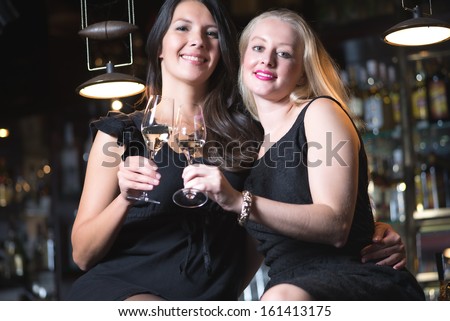 Two beautiful elegant women friends in stylish simple black cocktail dresses drinking a glass of chilled champagne as they celebrate on a night out together