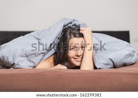 Smiling attractive young woman enjoying a lazy day lying on her stomach cuddling under a warm duvet and looking at the camera with a happy smile