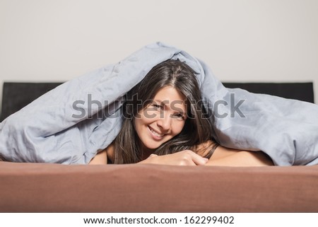 Smiling attractive young woman enjoying a lazy day lying on her stomach cuddling under a warm duvet and looking at the camera with a happy smile