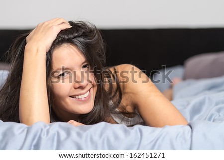 Smiling attractive young woman enjoying a lazy day lying on her stomach cuddling on a warm duvet and looking at the camera with a happy smile