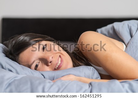 Smiling attractive young woman enjoying a lazy day lying on her stomach cuddling on a warm duvet and looking at the camera with a happy smile