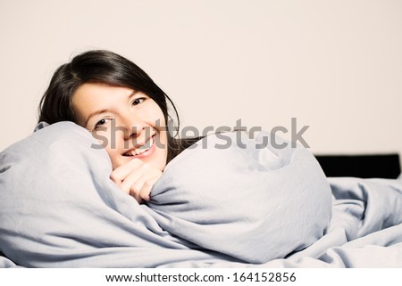 Laughing attractive young woman snuggling up in her comfortable warm bed peeking over the top of the duvet at the camera