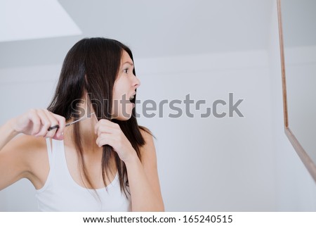 Attractive young woman with long brunette hair preparing to cut it with a pair of scissors in the bathroom grimacing in apprehension as she has misgivings about her decision