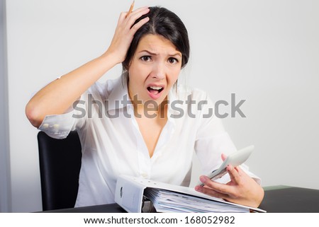 Businesswoman sitting at her desk with a large office binder looking at her calculator in horror or dismay with her mouth open in shock