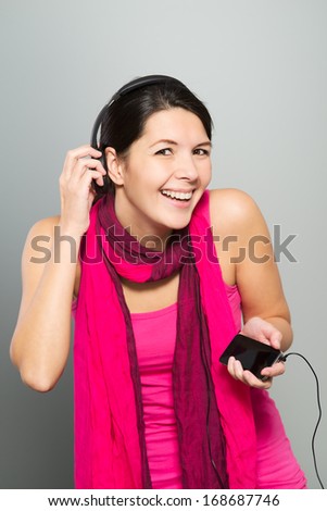 Beautiful vivacious woman listening to music on headphones connected to a handheld electronic storage device as she stands laughing at the camera