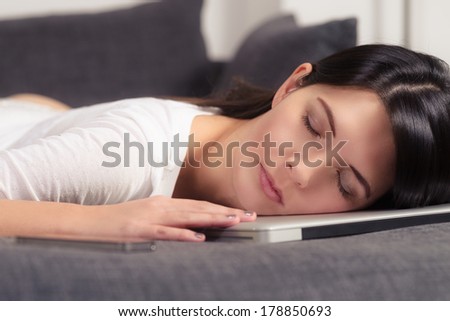 Attractive young woman resting her head on a laptop computer with her eyes closed, enjoying a peaceful sleep on the sofa