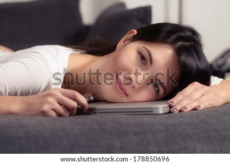 Attractive young woman resting her head on a laptop computer as she lays on the sofa taking a break and giving the camera a charming friendly smile