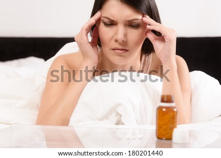 Woman with a migraine headache lying in bed frowning in pain with her hands to her temples contemplating the wisdom of taking a row of tablets laid out on a table in front of her