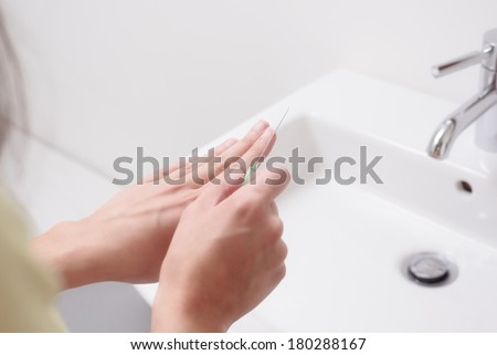 Young woman filing her finger nails with a metal file over a white ceramic hand basin in the bathroom in a nail care and beauty concept