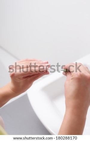 Young woman filing her finger nails with a metal file over a white ceramic hand basin in the bathroom in a nail care and beauty concept