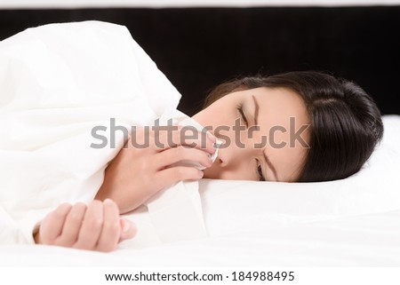Sick young woman lying in her clean white bed blowing her nose on a tissue conceptual of healthcare and seasonal flu and chills, rhinitis or an allergic reaction in hay fever