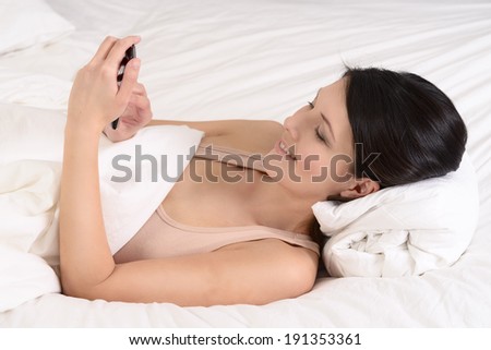 Attractive young woman lying in bed reading an sms on her mobile phone smiling with pleasure at the message, closeup side view