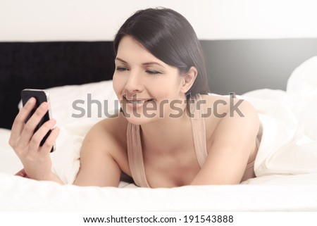 Attractive young woman lying in bed reading on sms on her mobile phone smiling with pleasure at the message, closeup side view