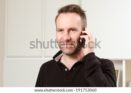 Horizontal portrait of a Caucasian middle-aged handsome man wearing a long-sleeved black T-shirt while having a pleasant conversation on the mobile phone, indoors