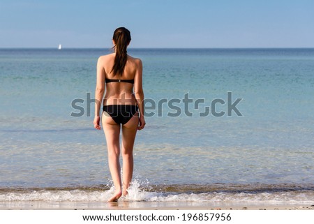Young woman with a lovely figure wearing a black bikini with her long brown hair tied in a ponytail walking away from the camera entering the sea on a hot summer day at the beach