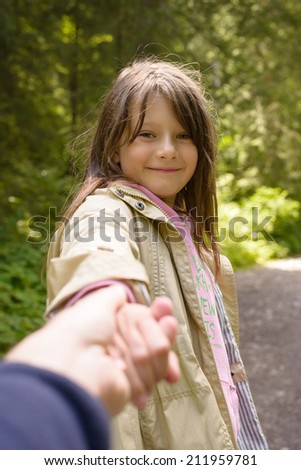 Beautiful young girl with a cute smile holding the hand of a parent turning back to look at the camera as they walk down a country lane with greenery