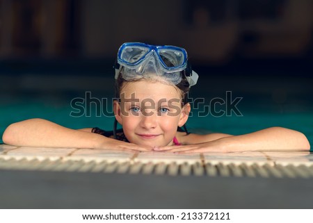 Pretty little girl wearing swimming goggles peering over the tiled edge of a swimming pool at the camera as she enjoys a relaxing day of playing in the water during her summer vacation