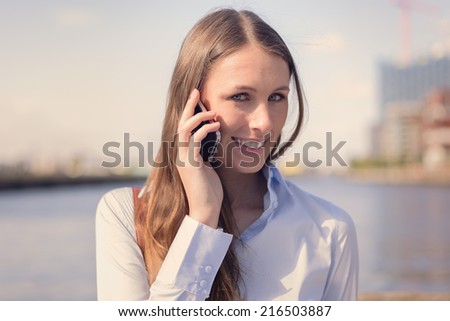 Attractive woman taking a call on her mobile phone standing outdoors against a river background smiling at the camera as she listens to the conversation