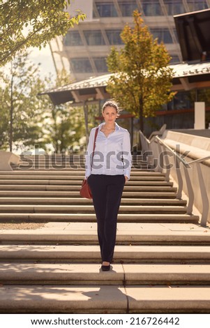 Slender elegant young woman carrying a handbag descending a flight of open-air concrete stairs in town with modern commercial buildings behind her