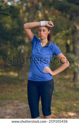 Woman athlete wiping sweat from her forehead onto her wristband as she pauses during her training exercises on a forest track