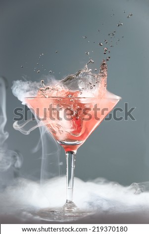 Smoking martini cocktail in a conical glass with wafting vapor and splashing droplets from a falling cherry for a dramatic effect over a grey background