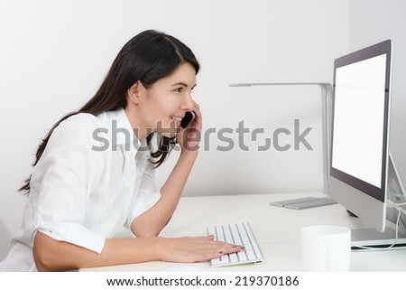 Businesswoman working in her office sitting at a desk in front of her desktop computer with a blank screen visible to the viewer as she chats on her mobile phone, side view