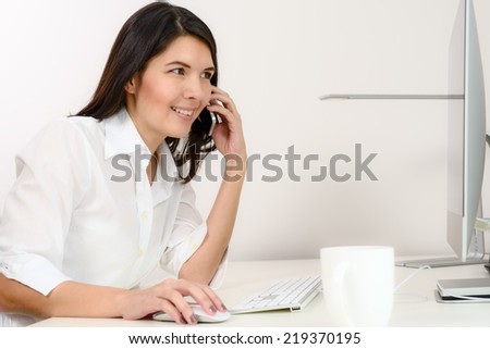 Businesswoman working in her office sitting at a desk in front of her desktop computer as she chats on her mobile phone, side view